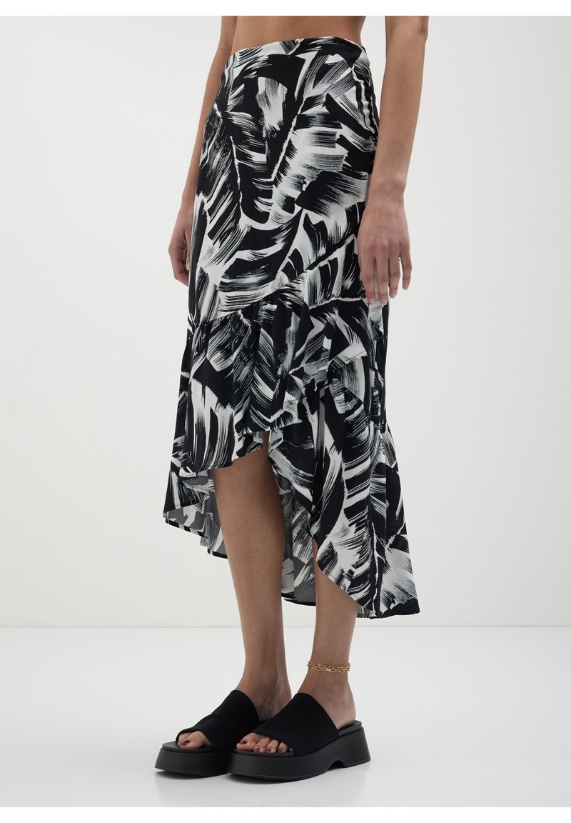 Jupe midi à imprimé abstrait noir et blanc avec un ourlet asymétrique, portée avec des sandales slides à plateforme noires et une chaîne de cheville dorée sur fond clair.