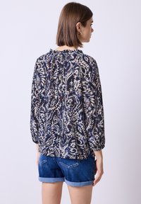 Langärmlige Bluse in Marineblau mit einem Blumenmuster in Creme und Rot. Verfügt über einen gerafften Kragen und einer lockeren Passform. Kombiniert mit Jeansshorts.