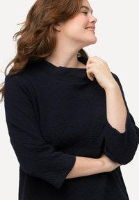 Top blu navy con finitura testurizzata, caratterizzato da un collo alto e maniche a tre quarti. Tessuto morbido con un delicato motivo.