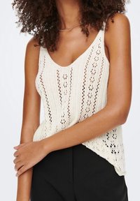 Top in maglia di colore crema con una texture leggera, caratterizzato da motivi a traforo e spalline regolabili, abbinato a pantaloni neri.