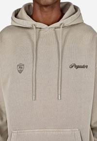 Bēša kapuccīna ar priekšu kabatu, pavilkšanas auklām un sudraba agletiem. Iekļauj logo un teksta izšuvumu kreisajā krūšu daļā.