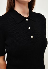 Haut en maille côtelée noire avec un col, doté de trois boutons métalliques et de manches courtes. Le tissu texturé donne une apparence ajustée.