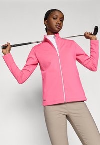 Pink zip-up jacket made of soft material with a high collar and two side pockets. Model poses with a golf club, wearing beige pants.