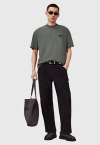 Camiseta de algodón verde oscuro con mangas cortas, combinada con pantalones de pana negros, zapatos oscuros y una bolsa de tela gris.