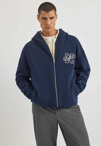 Granatowa bluza z kapturem zapinana na zamek, wykonana z miękkiego materiału, z kieszenią kangurką oraz haftowanym białym logo. Model nosi pod spodem szare spodnie.