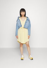 Vestido amarelo com decote em V e cintura com cordão, sobreposto por uma jaqueta de ganga curta. Usado com ténis brancos e amarelos.