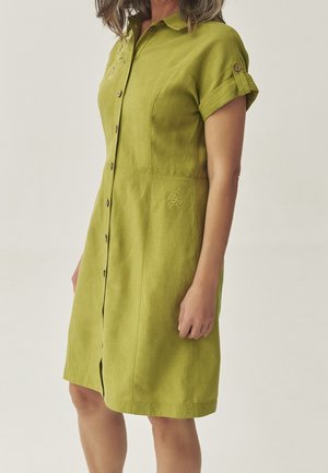 Donna che indossa un vestito verde oliva, corto, a maniche corte e con bottoni, lungo fino al ginocchio, con dettagli ricamati e bottoni in legno, in piedi davanti a uno sfondo neutro.