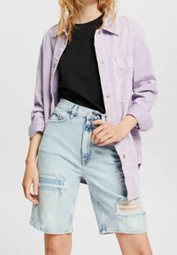 Femme portant une chemise lilas clair ouverte sur un top noir et un short en jean bleu clair déchiré.