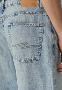 Jean en denim bleu clair avec une poche arrière ornée d'un détail de surpiqure en zigzag et d'une étiquette en cuir marron portant l'inscription "QS."