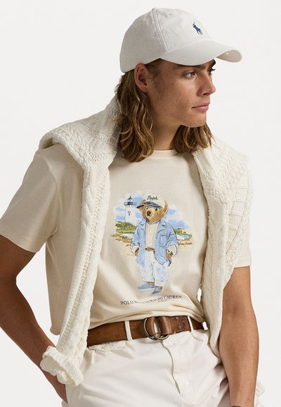 Kortærmet beige t-shirt med et bjørnegrafik i denimoutfit, draperet med en hvid strikket sweater, parret med lyse bukser og en kasket.