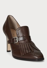 Brown leather high heel shoe with fringe detail, brogue perforations, a buckle strap, and a pointed toe design. Smooth texture throughout.