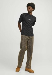 Zwarte t-shirt met korte mouwen en witte tekst, gecombineerd met losse bruine gestreepte jeans en lichtbruine werkschoenen. Model staat met handen in zijn zakken.