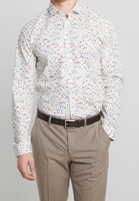 Camisa blanca de botones con un patrón floral en rojo, azul y verde, hecha de una tela ligera. Combinada con pantalones beiges y un cinturón marrón.
