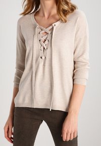 Beige sweater with a V-neck and lace-up front. Long sleeves, ribbed cuffs, and soft texture. Paired with brown textured pants.