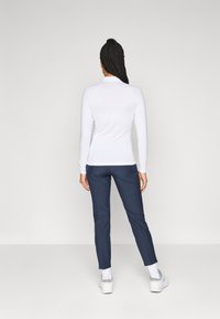Femeie cu păr împletit lung stând cu spatele, purtând un top alb cu mâneci lungi, pantaloni albastru închis, șosete albe și adidași albi.