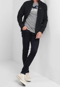 Černá barva košile s knoflíky přes šedé tričko Hollister, doplněná o tmavé džíny a bílé tenisky. Oblečení má neformální, vrstvený vzhled.