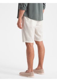 Shorts beige clair, arrivant au genou, avec une coupe décontractée et une légère texture, associés à des chaussures slip-on beiges, et une chemise vert foncé.