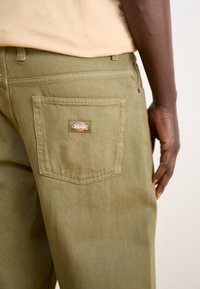Olive green pants made of durable fabric, featuring a back pocket with a branded patch and visible stitching details.
