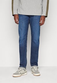 Blue denim jeans with a straight-leg cut, featuring a medium wash, contrasting stitching, and worn with gray athletic shoes.