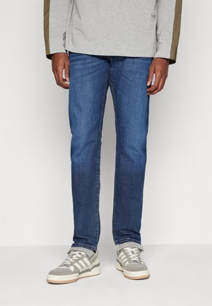 Blue denim jeans with a straight-leg cut, featuring a medium wash, contrasting stitching, and worn with gray athletic shoes.