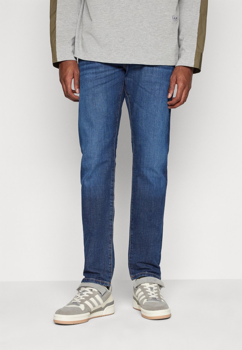 Blue denim jeans with a straight-leg cut, featuring a medium wash, contrasting stitching, and worn with gray athletic shoes.