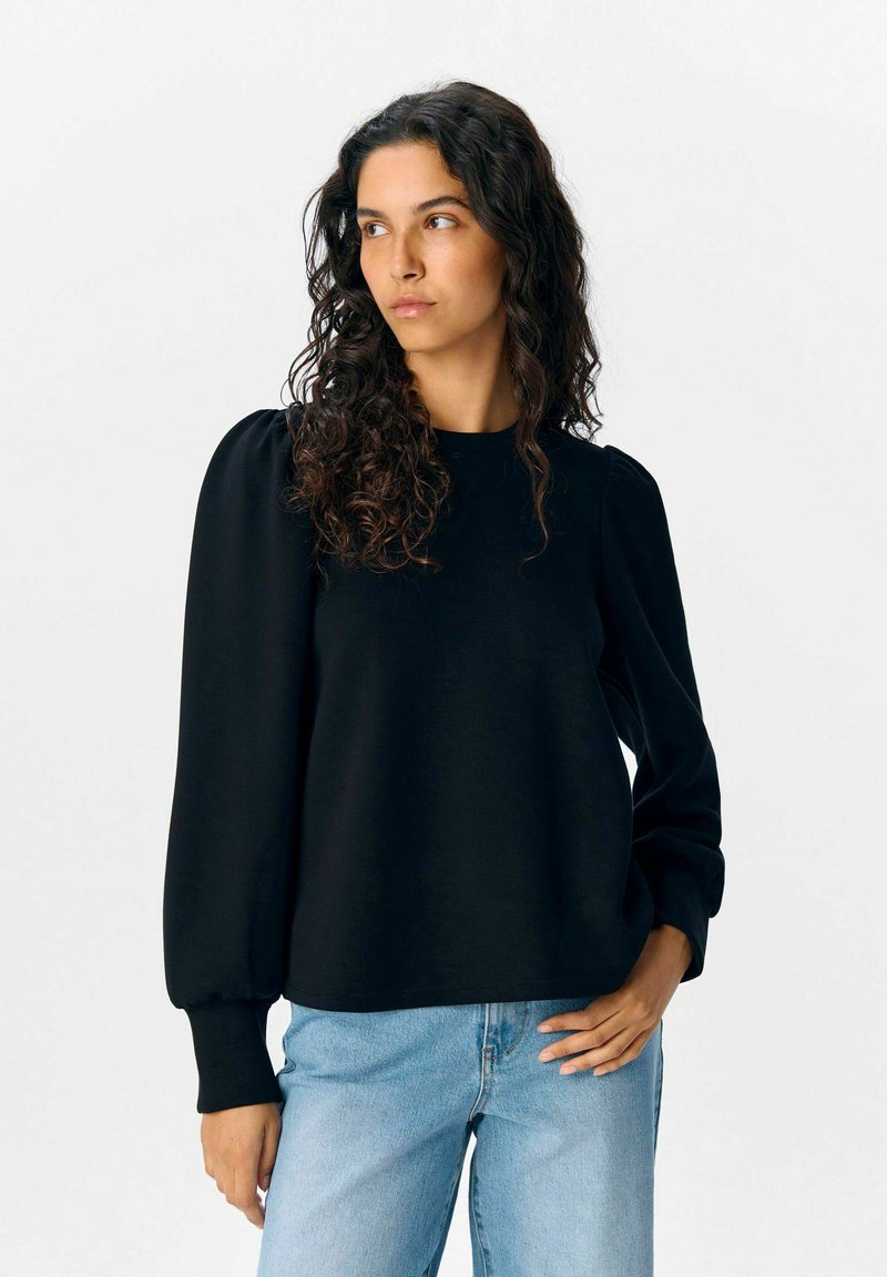 Sweatshirt noir à manches longues et bouffantes avec un col rond. Texture douce, associé à un jean droit bleu clair.