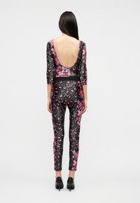 Γυναίκα όρθια πλάγια, φοράει ολόσωμη φόρμα ροζ και μαύρου χρώματος με βαθύ ντεκολτέ στην πλάτη και μαύρες ψηλοτάκουνες γόβες.