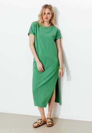 Femme portant une robe midi verte fendue sur le côté, des bracelets dorés, des colliers superposés et des sandales imprimé léopard, debout contre un mur blanc.