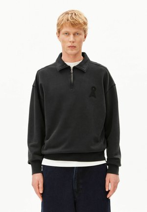 Blonder Mann trägt einen schwarzen Pullover mit Viertel-Reißverschluss und kleinem schwarzem Logo auf der linken Brust, weißes Unterhemd und dunkelblaue Hose vor weißem Hintergrund.