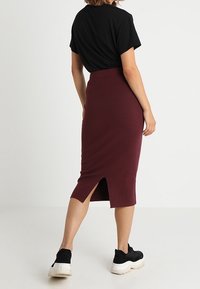 Schwarzes kurzärmliges Oberteil, kombiniert mit einem figurbetonten burgunderfarbenen Midirock mit einem Schlitz hinten. Das Model trägt schwarze Sneaker. Einfaches und elegantes Design.