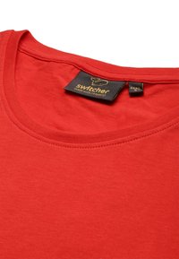 T-shirt rossa in cotone con scollo rotondo, con un'etichetta che recita "switcher" in oro su uno sfondo nero. Texture liscia.