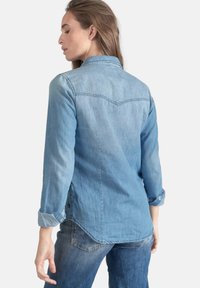 Chemise en denim bleu clair à coupe droite, avec des poignets à bouton, un empiècement au dos et un ourlet légèrement arrondi. Texture douce et design décontracté.