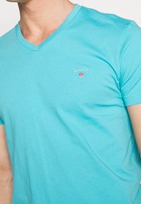 Türkises Baumwoll-T-Shirt mit V-Ausschnitt. Mit einem kleinen gestickten Logo auf der Brust. Weicher Griff und kurze Ärmel.