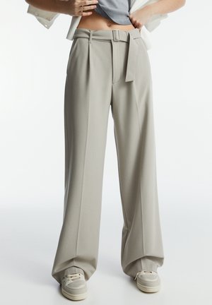 Personne portant un pantalon beige taille haute à jambes larges avec une ceinture assortie et des baskets blanches, levant un haut gris pour révéler le ventre.
