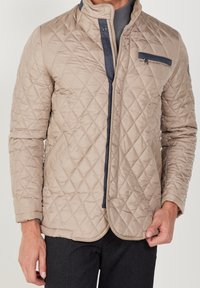 Beige quiltad jacka med hög krage, dragkedja fram och blå accentficka. Texturerat tyg och slätt foder syns.