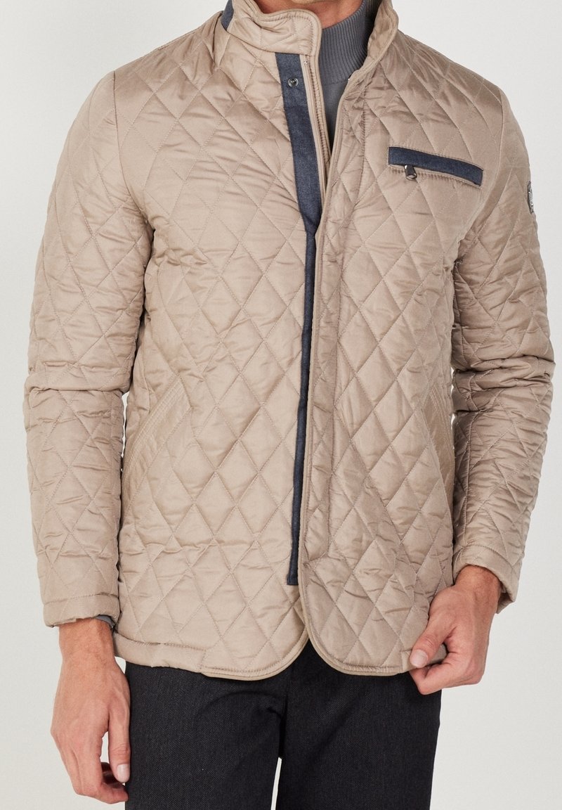 Beige quiltet jakke med høj krave, lynlås foran og blå accentlomme. Tekstureret stof og glat foring synlig.