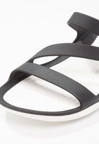 Sandales noires avec deux larges sangles croisées en matériau doux, dotées d'une semelle blanche flexible avec un motif texturé.
