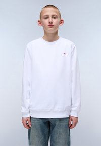 Hvit genser med lange ermer og ribbestrikkede mansjetter, med en liten rød og blå logo på brystet, sammen med lyseblå jeans.