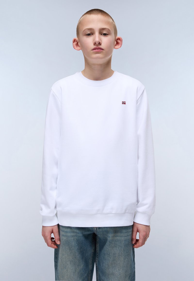 Hvit genser med lange ermer og ribbestrikkede mansjetter, med en liten rød og blå logo på brystet, sammen med lyseblå jeans.