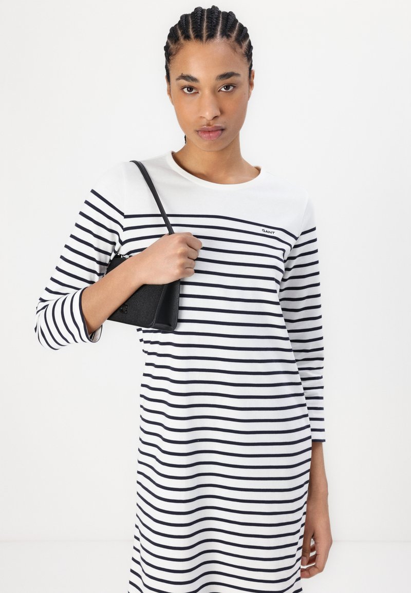 Pasiasta sukienka z długim rękawem w białym i granatowym kolorze. Wygodny materiał bawełniany z okrągłym dekoltem. W jednej ręce trzymana jest mała czarna torebka.