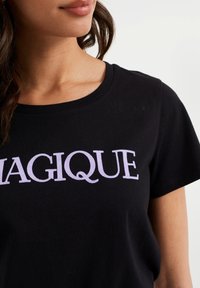 Černé bavlněné tričko s krátkými rukávy a kulatým výstřihem, s fialovým blokovým písmem, které přes hrudník hlásí "MAGIQUE".