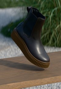 Bruine leren enkelboots met een ribbelrubberen zool, elastische zijpanelen en een ronde neus, tentoongesteld op een houten oppervlak.