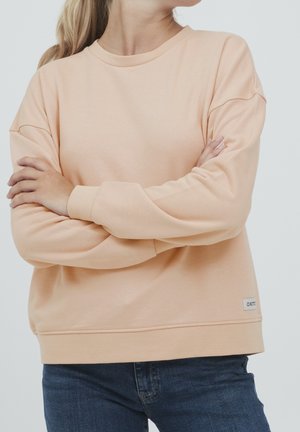 Osoba w jasnoróżowej bluzie i ciemnoniebieskich dżinsach, z założonymi skrzyżowanymi rękami, z długimi włosami związanymi do tyłu, na jednolitym tle.