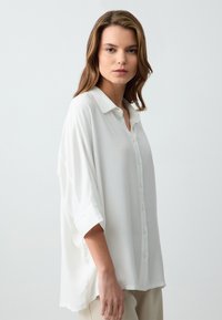 Weiße, locker sitzende Bluse mit einem Kragen, geknöpftem Vorderteil und hochgerollten kurzen Ärmeln, aus leichtem Stoff mit einer glatten Textur.
