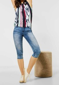 Kvinna klädd i en färgglad randig ärmlös topp, avklippta blå jeans och vita sneakers, stående bredvid en beige texturerad rund pall.