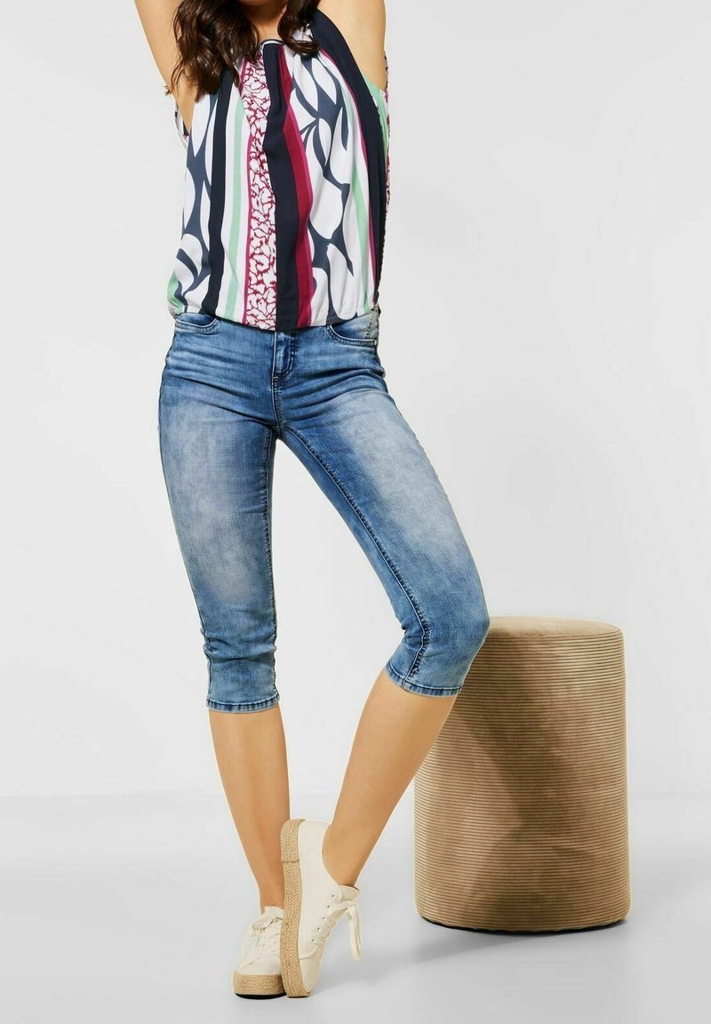 Kvinna klädd i en färgglad randig ärmlös topp, avklippta blå jeans och vita sneakers, stående bredvid en beige texturerad rund pall.