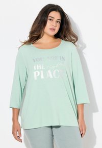 Mátově zelený top s dlouhými rukávy a kulatým výstřihem, na přední straně s metalicky stříbrným nápisem "JSTE NA SPRÁVNÉM MÍSTĚ".