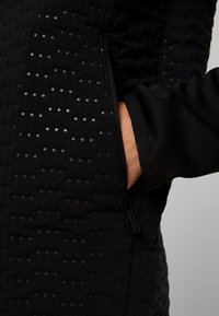 Chaqueta aislante negra con un patrón de rombos y puntos negros, que cuenta con un bolsillo lateral con cremallera y una tela texturizada.