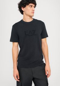 T-shirt preto de manga curta com acabamento texturizado e impressão do logótipo à frente. Combinado com calções cargo escuros, de design minimalista.