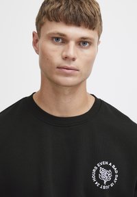 Svarte hettegensere med rund hals, med hvit blomsterdesign og tekst. Myk tekstur, standard passform, synlige ribbestrikkede mansjetter.